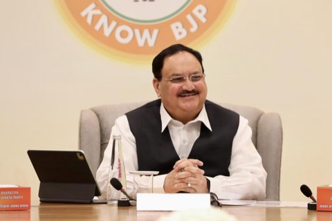 Jagat Prakash Nadda seated at a table, smiling, with a Know BJP emblem on the wall behind