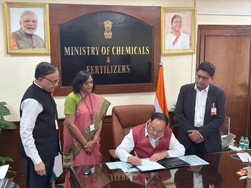 Jagat Prakash Nadda signing papers at a desk beneath the Ministry of Chemicals and Fertilizers sign, officials standing alongside