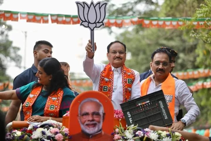 Jagat Prakash Nadda standing on a vehicle during a roadshow, raising a party symbol